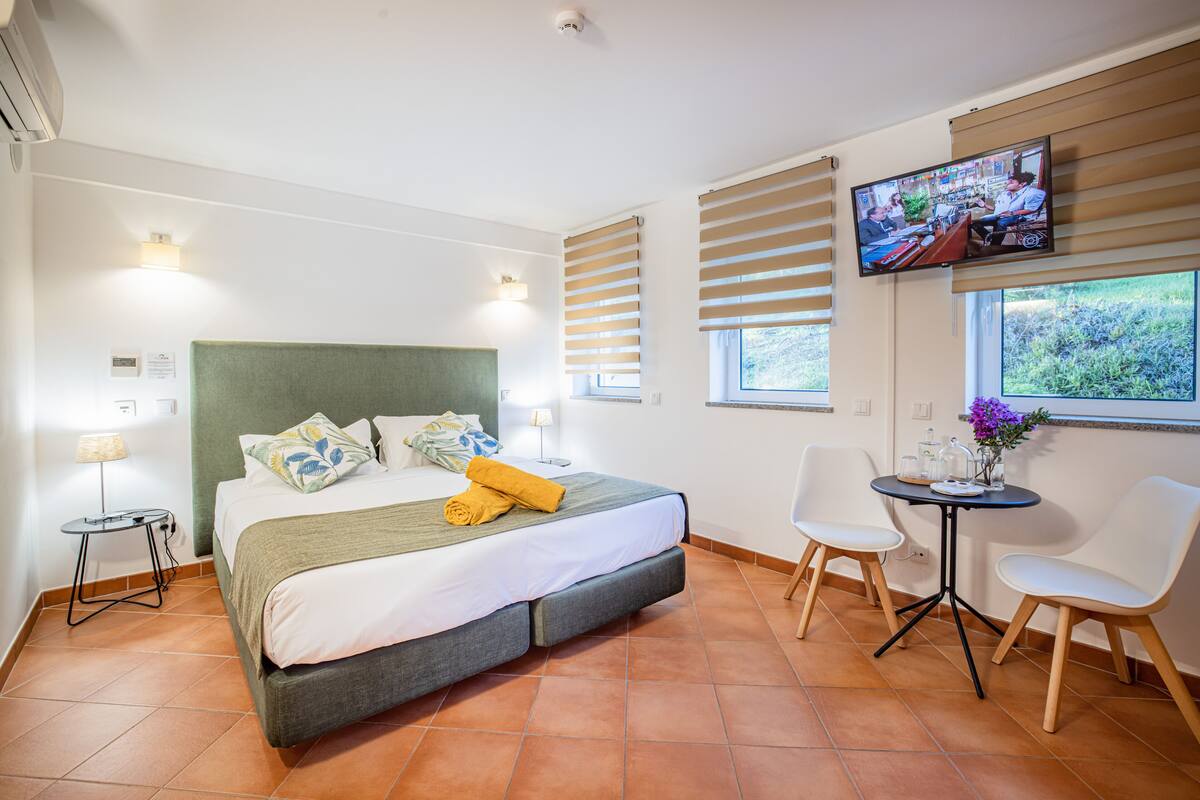 A modern hotel room with a large bed, a television, a small table with a vase, and a window with blinds.