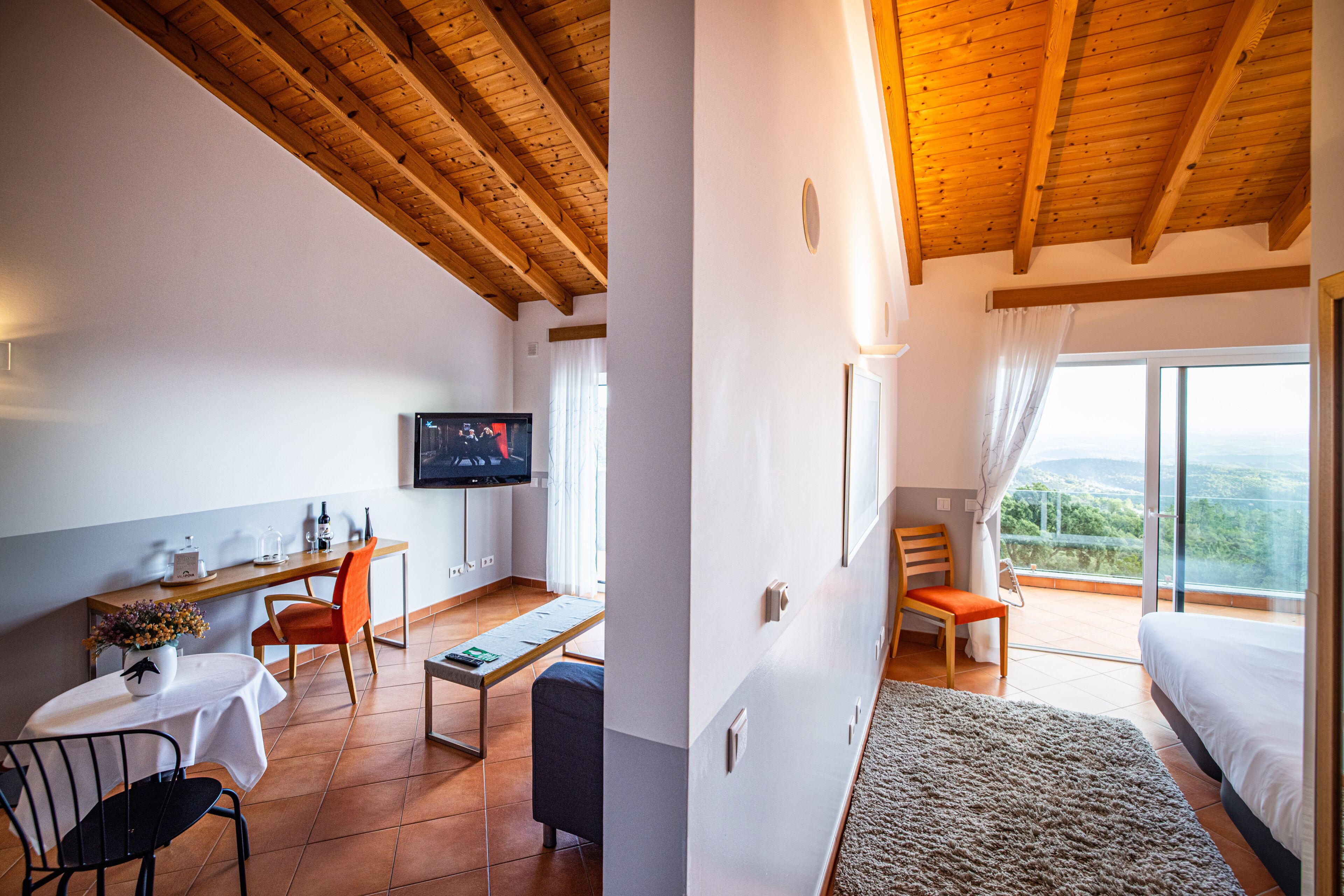 A room with a wooden ceiling, a TV on the wall, a desk with a chair, a bed, and a balcony with a view.