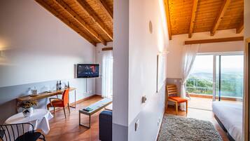 A room with a wooden ceiling, a TV on the wall, a desk with a chair, a bed, and a balcony with a view.