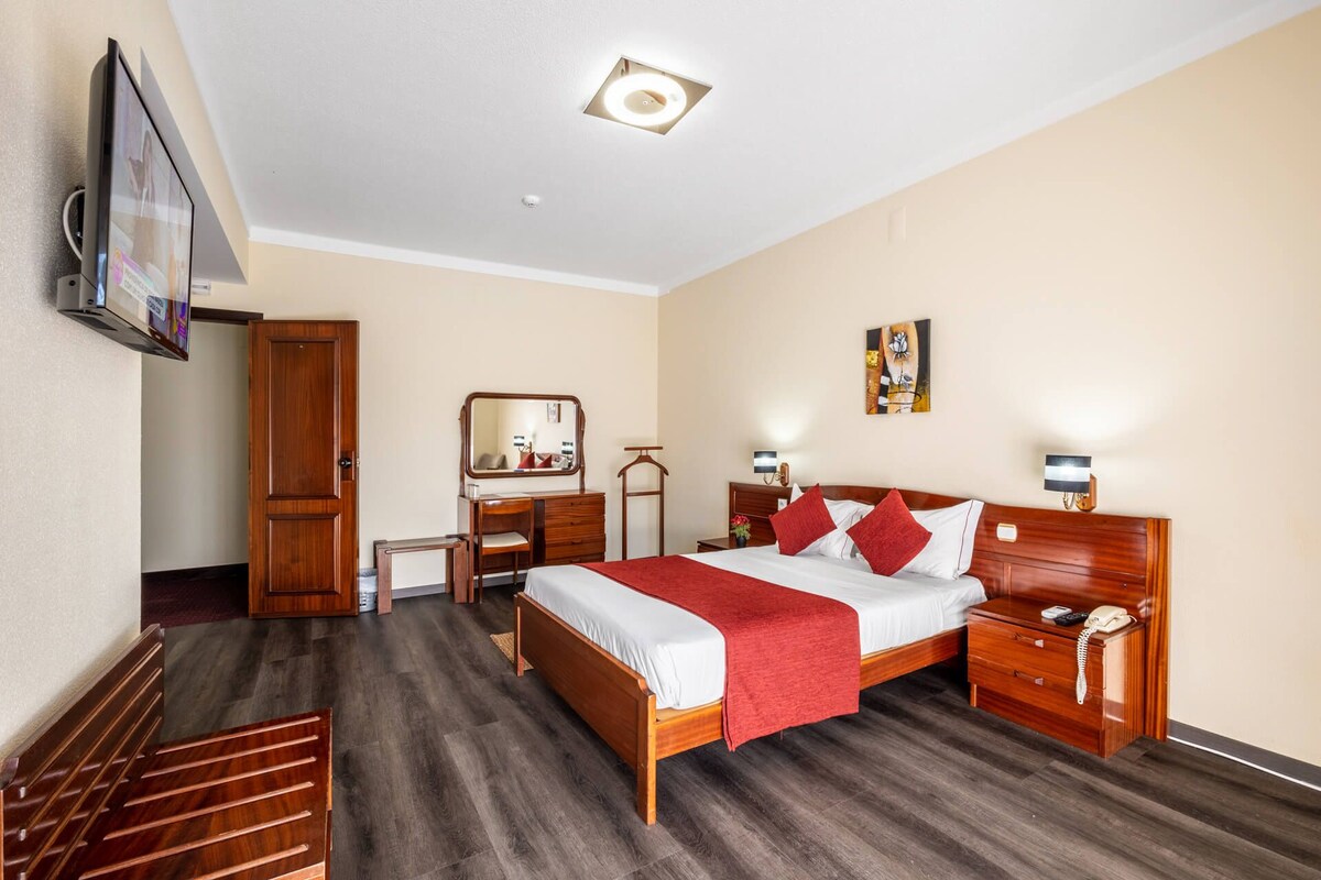 A hotel room with a wooden bed, bedside tables, a desk, a mirror, a TV, and a ceiling light.