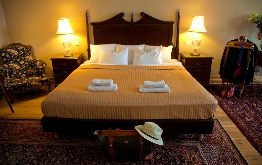 Cama bem arrumada com lençóis brancos e duas toalhas dobradas. Há duas mesas de cabeceira com luminárias, uma cadeira com estofamento estampado e um enfeite pendurado na parede.