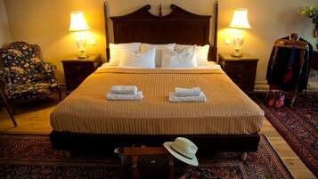 A neatly made bed with white linens and two towels folded on it. There are two bedside tables with lamps, a chair with a patterned upholstery, and a decorative wall hanging.