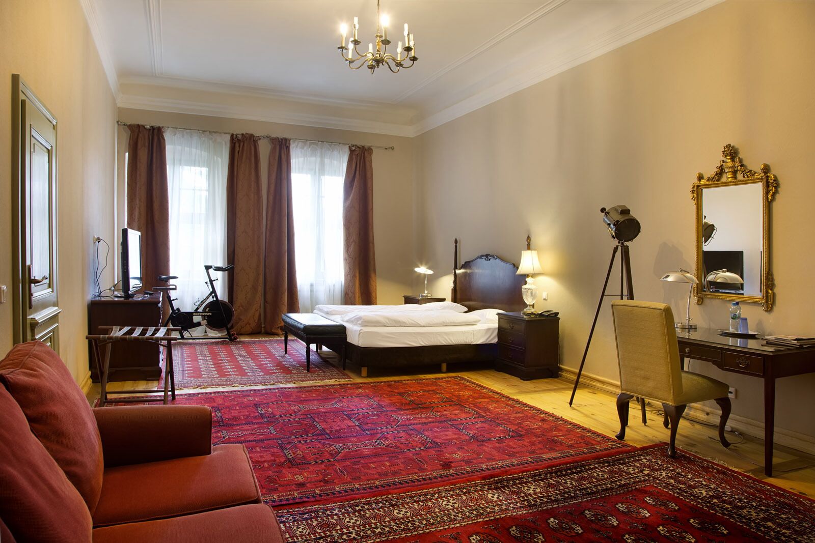 A hotel room with a bed, a desk with a chair, a television, a bicycle, and a large red carpet.
