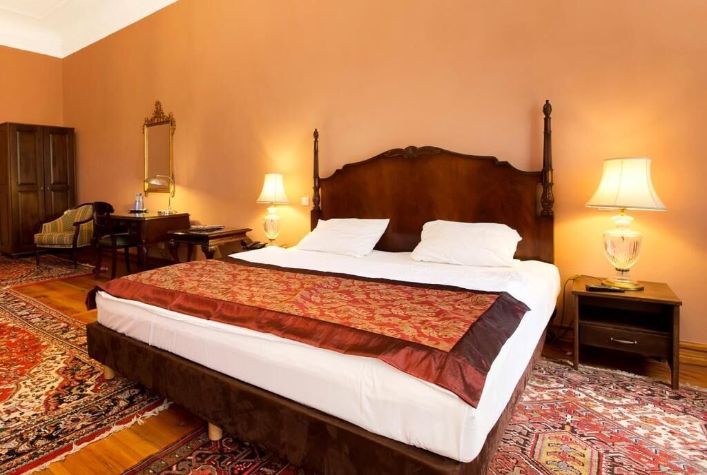 Habitación de hotel con una cama grande, dos mesitas de noche con lámparas, un armario de madera, un escritorio con silla y una alfombra estampada.
