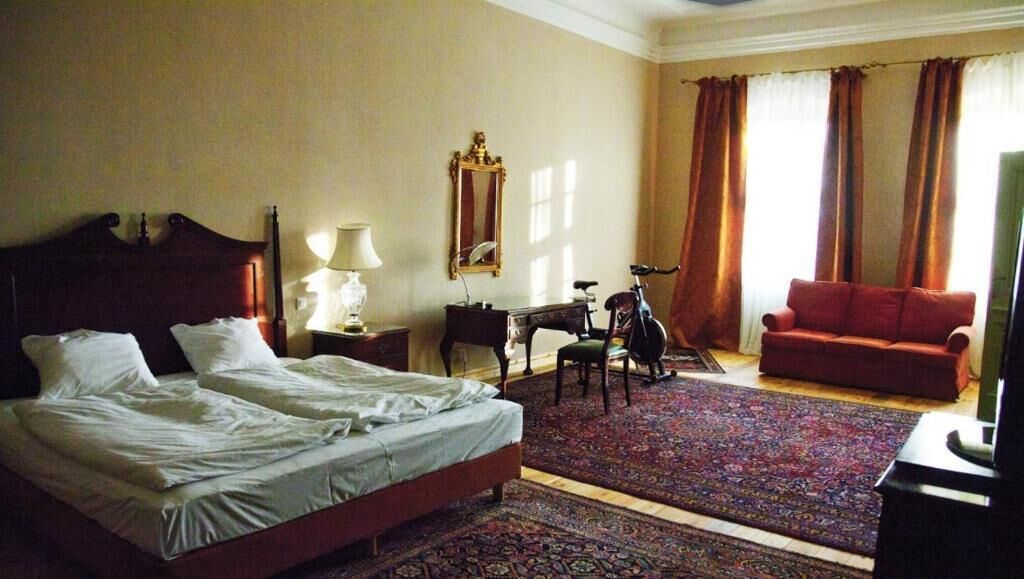 A hotel room with a large bed, a desk with a lamp, a chair, a red sofa, and a television.