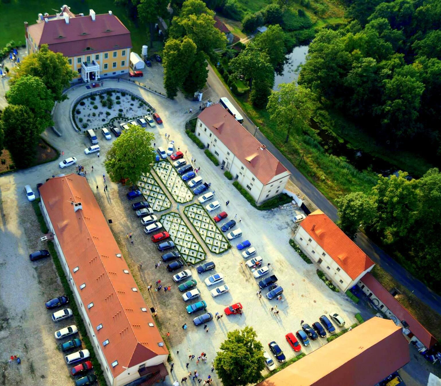 Eine Vogelperspektive auf einen Parkplatz mit mehreren Autos, einer begrünten Mittelinsel und Gebäuden mit roten Dächern.