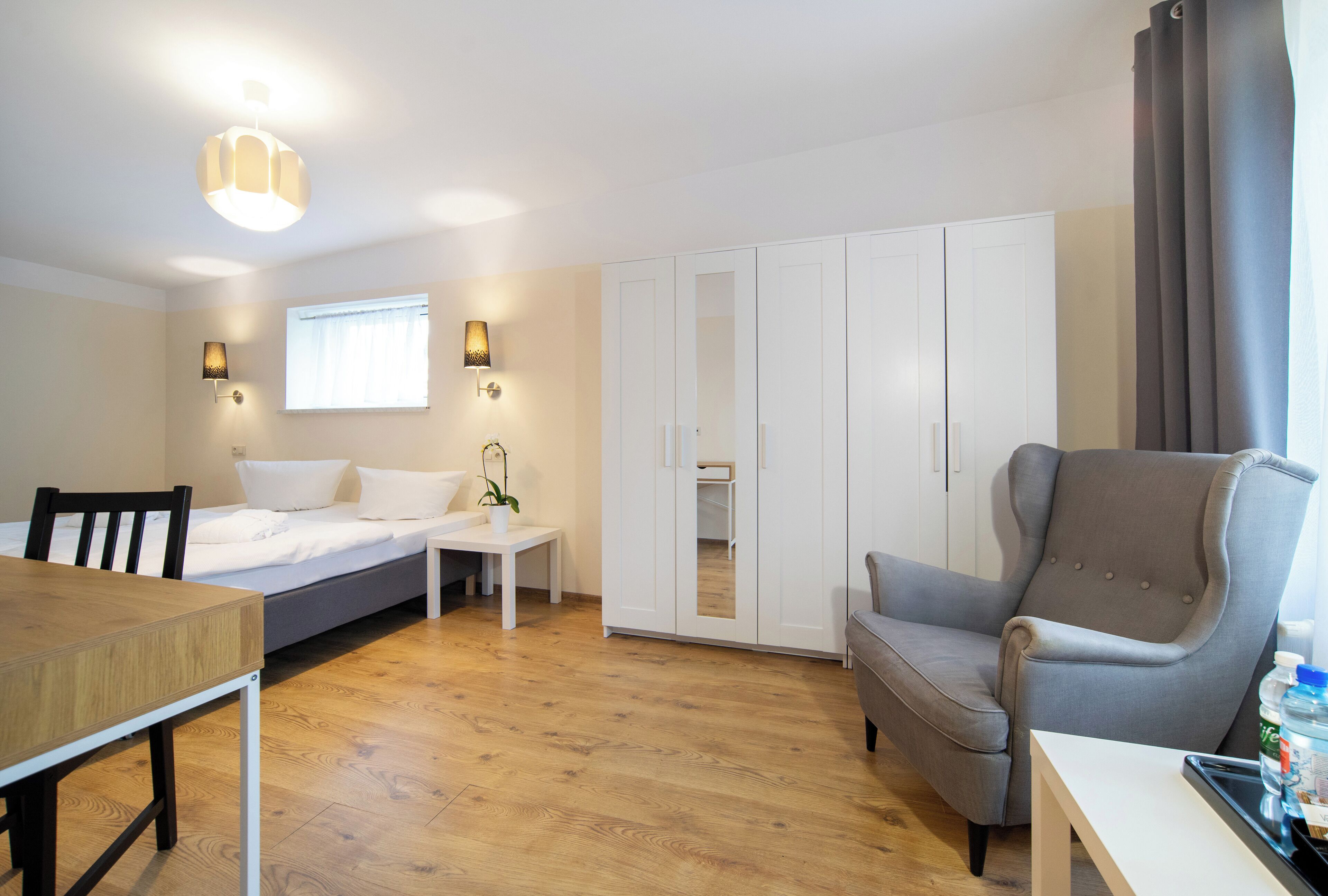 Quarto moderno com cama, mesa de cabeceira, abajur, guarda-roupa e uma poltrona cinza.