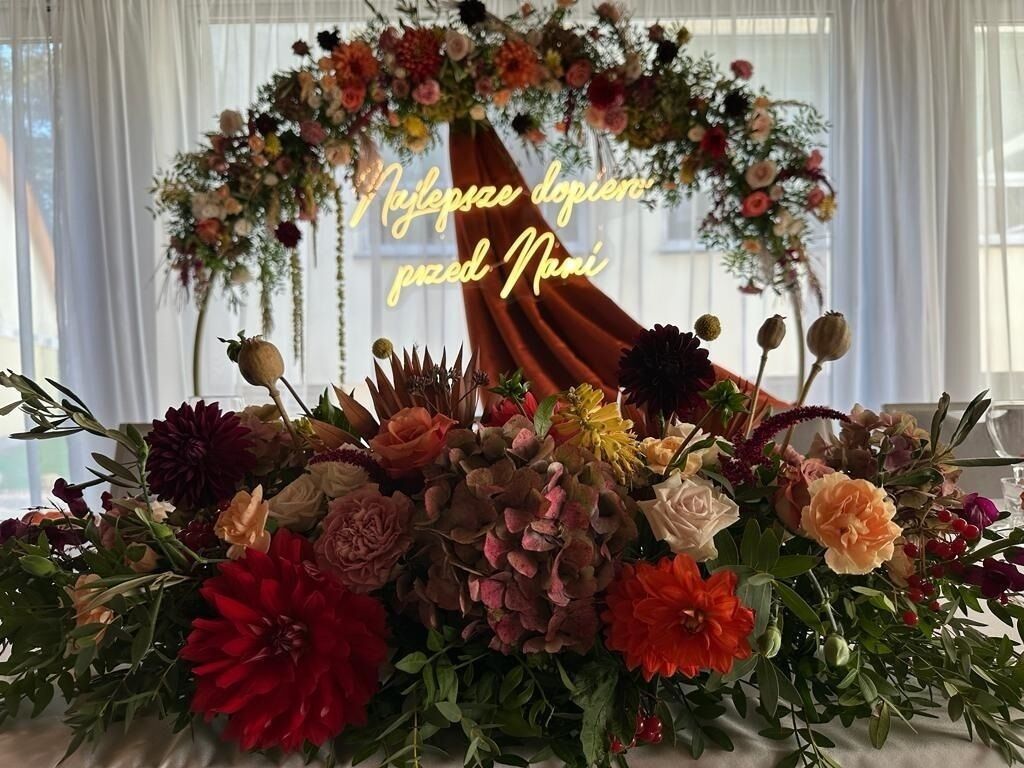 A floral arrangement with a variety of flowers and greenery, set against a backdrop of white curtains and a window.