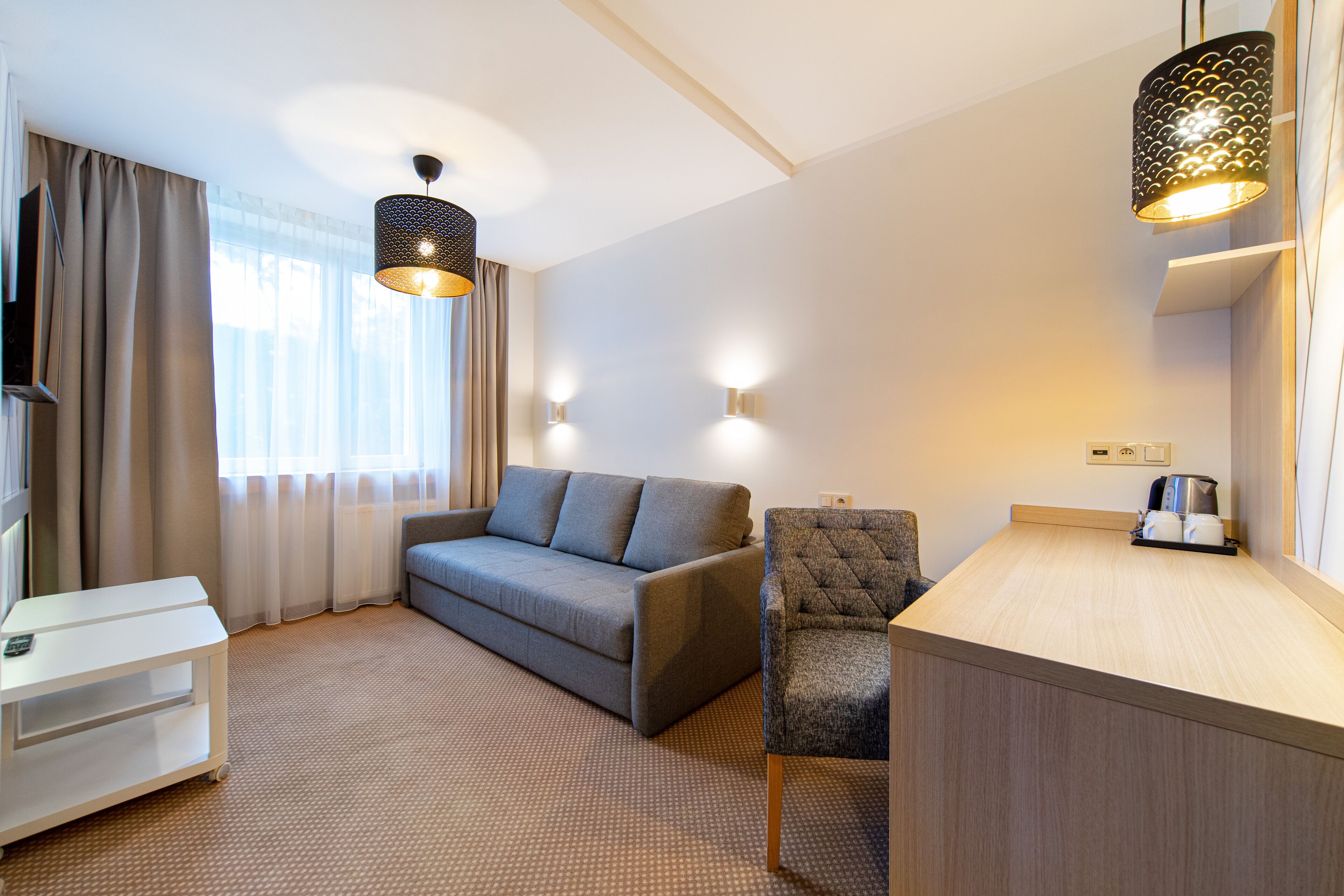 Ein modernes Hotelzimmer mit grauem Sofa, einem kleinen Sessel, einer weißen TV-Konsole und einem hellen Holztisch.