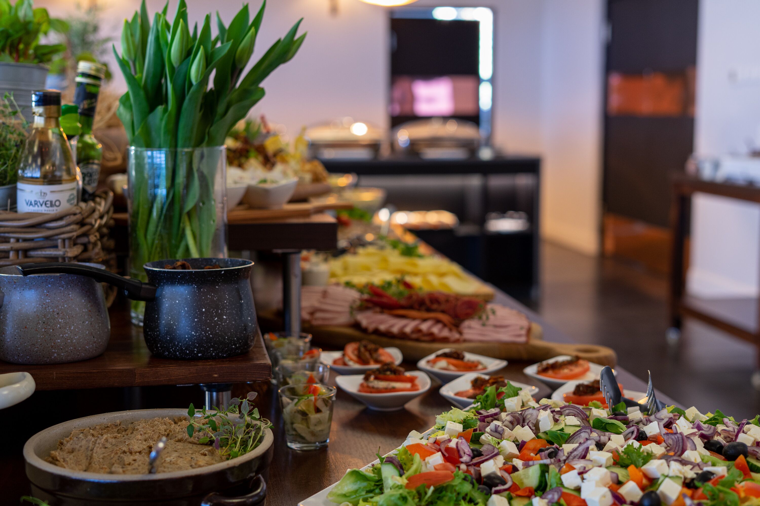 A buffet table with various dishes, including salads, meats, and cheeses. There are also some decorative items like a vase with green leaves and a bottle labeled "VARVELO".