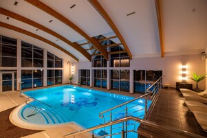 Una piscina interior con vigas de madera, ventanas amplias y una zona de estar junto a la piscina.