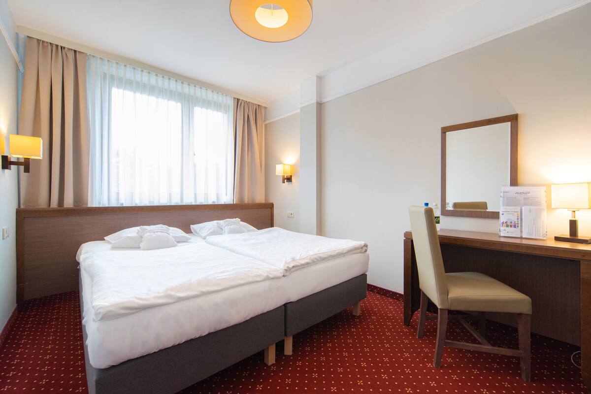 A hotel room with a double bed, a desk with a chair, a desk lamp, and a large mirror.