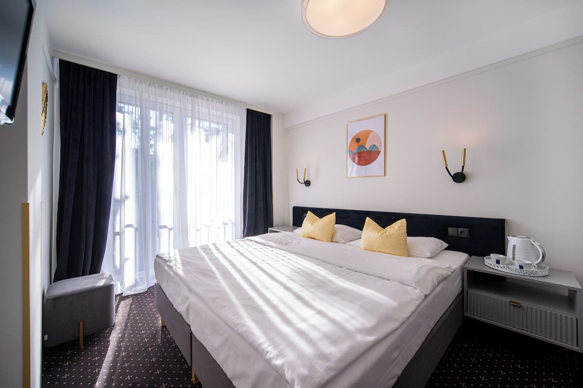 A hotel room with a large bed, a bedside table with a kettle, and a window with curtains.