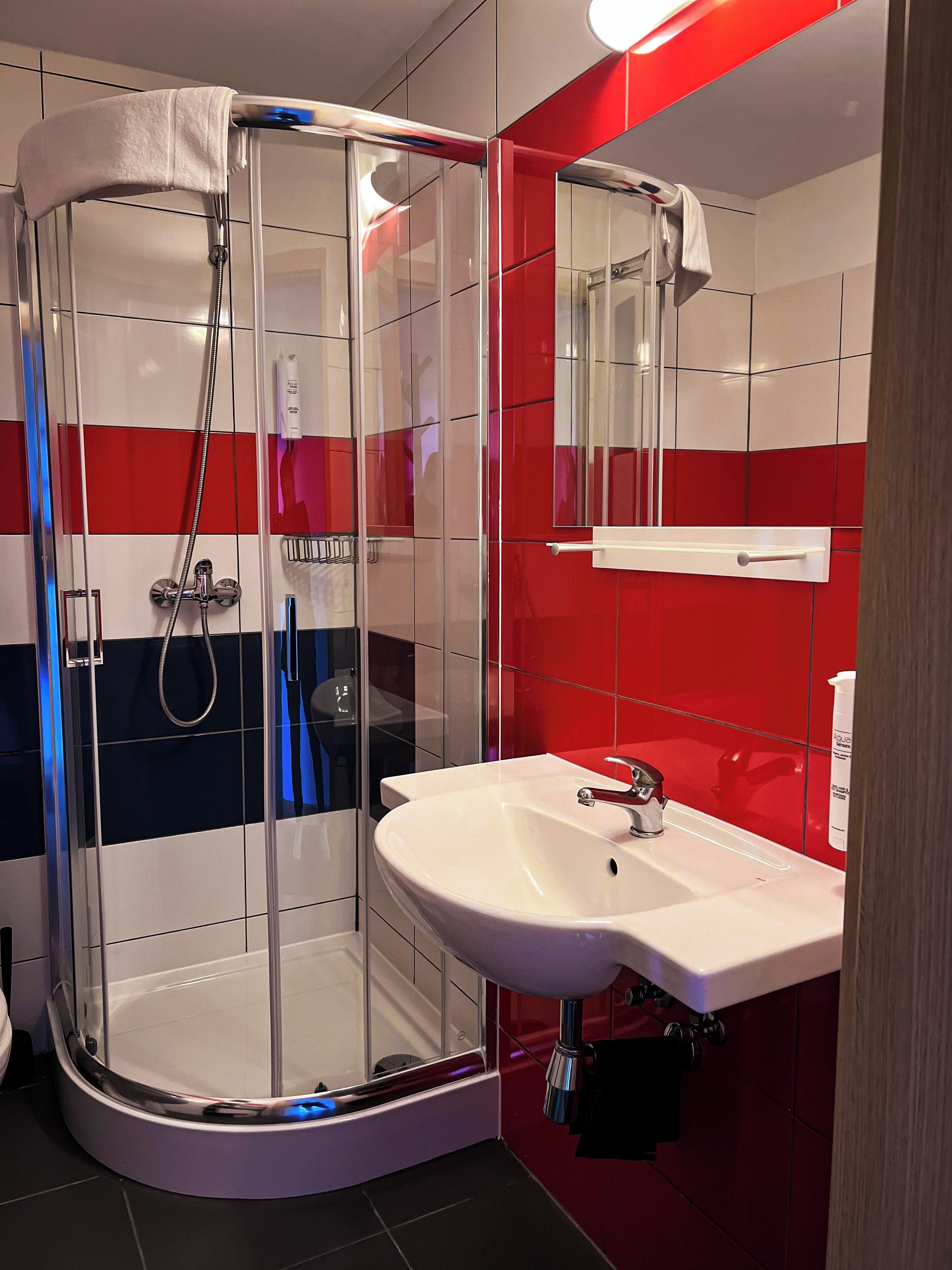 Un baño moderno con ducha envuelta en cristal, un lavabo blanco y paredes con azulejos rojos y blancos.