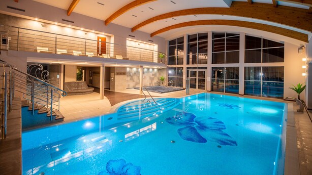 Piscina interior com teto de madeira e paredes de vidro, rodeada por uma área de estar e uma escadaria.