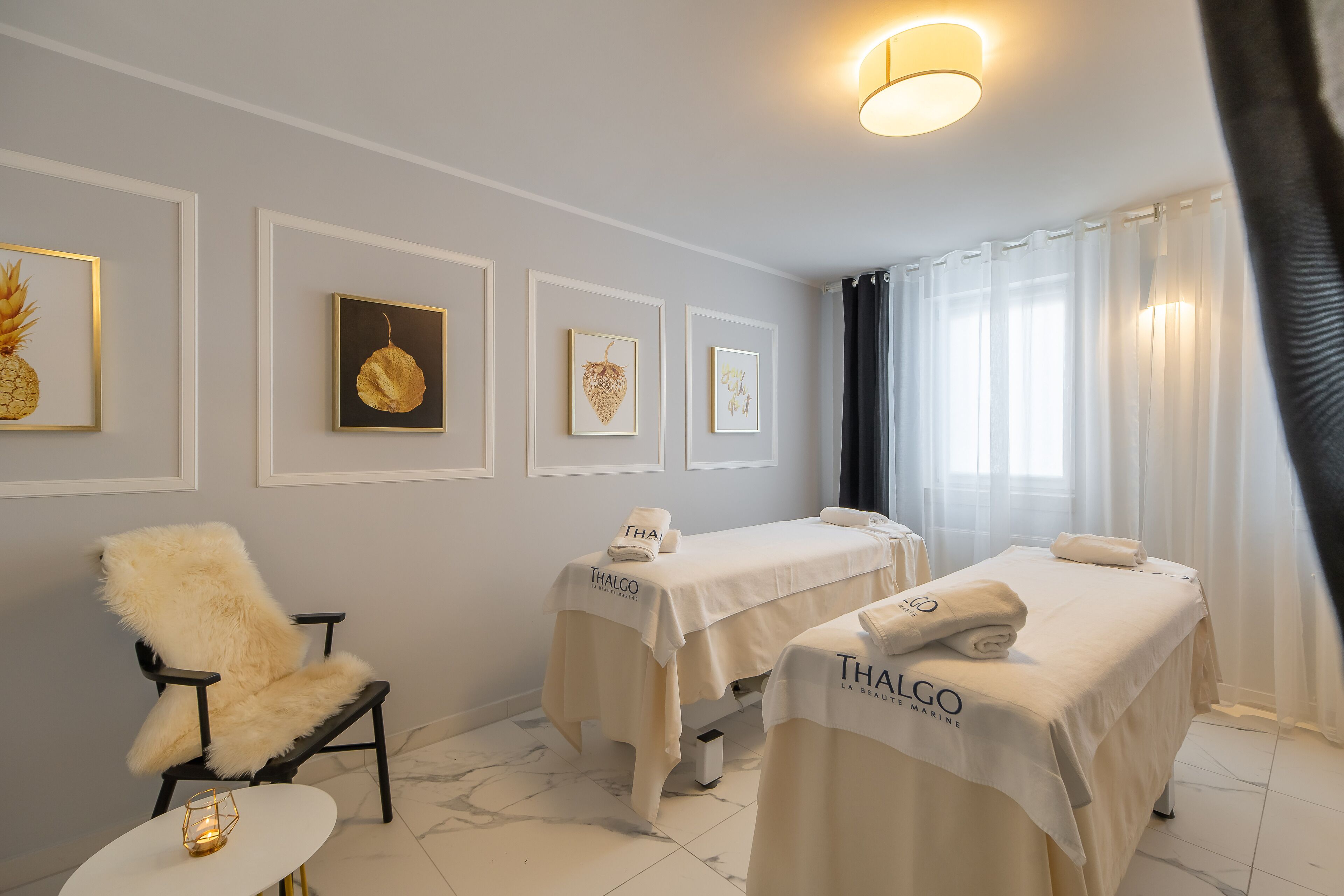 Egy wellness-szoba, melyben két masszázságy, egy puha fotel és a falon elhelyezett, keretezett műalkotások találhatók.