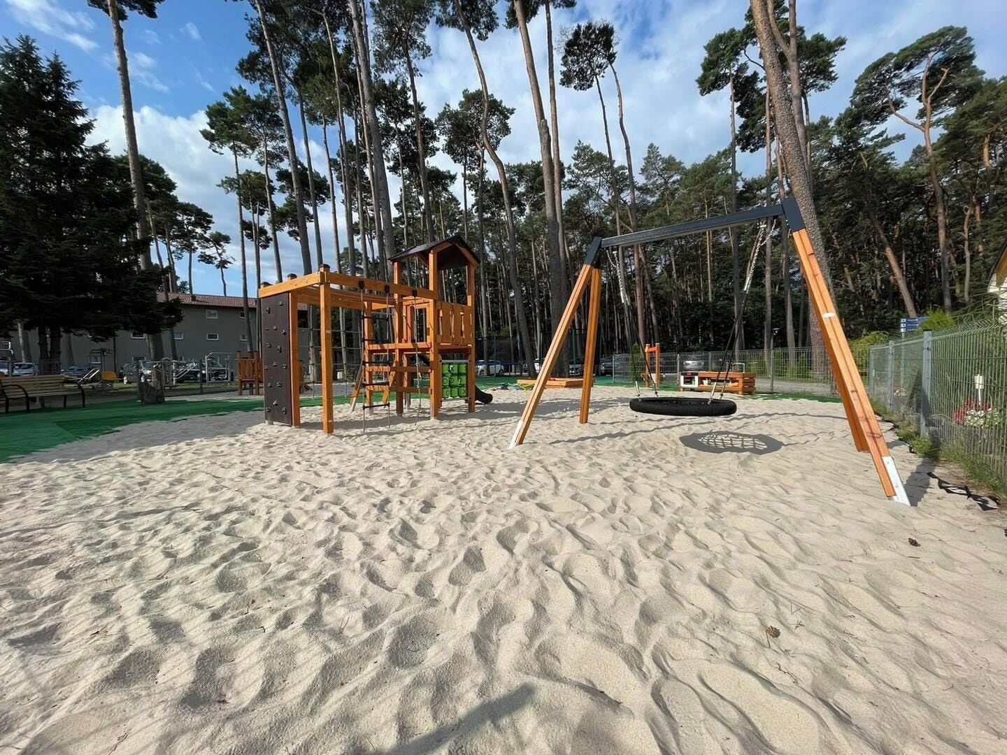 Une aire de jeux avec une balançoire en bois et une petite maison de jeu, le tout installé sur une zone sablonneuse entourée d’arbres.