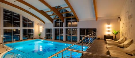 Ein Hallenbad mit hoher Decke und Holzbalken, umgeben von Liegestühlen und großen Fenstern.