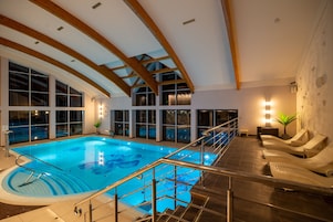 Una piscina interior con techo alto y vigas de madera, rodeada de sillas de descanso y grandes ventanales.
