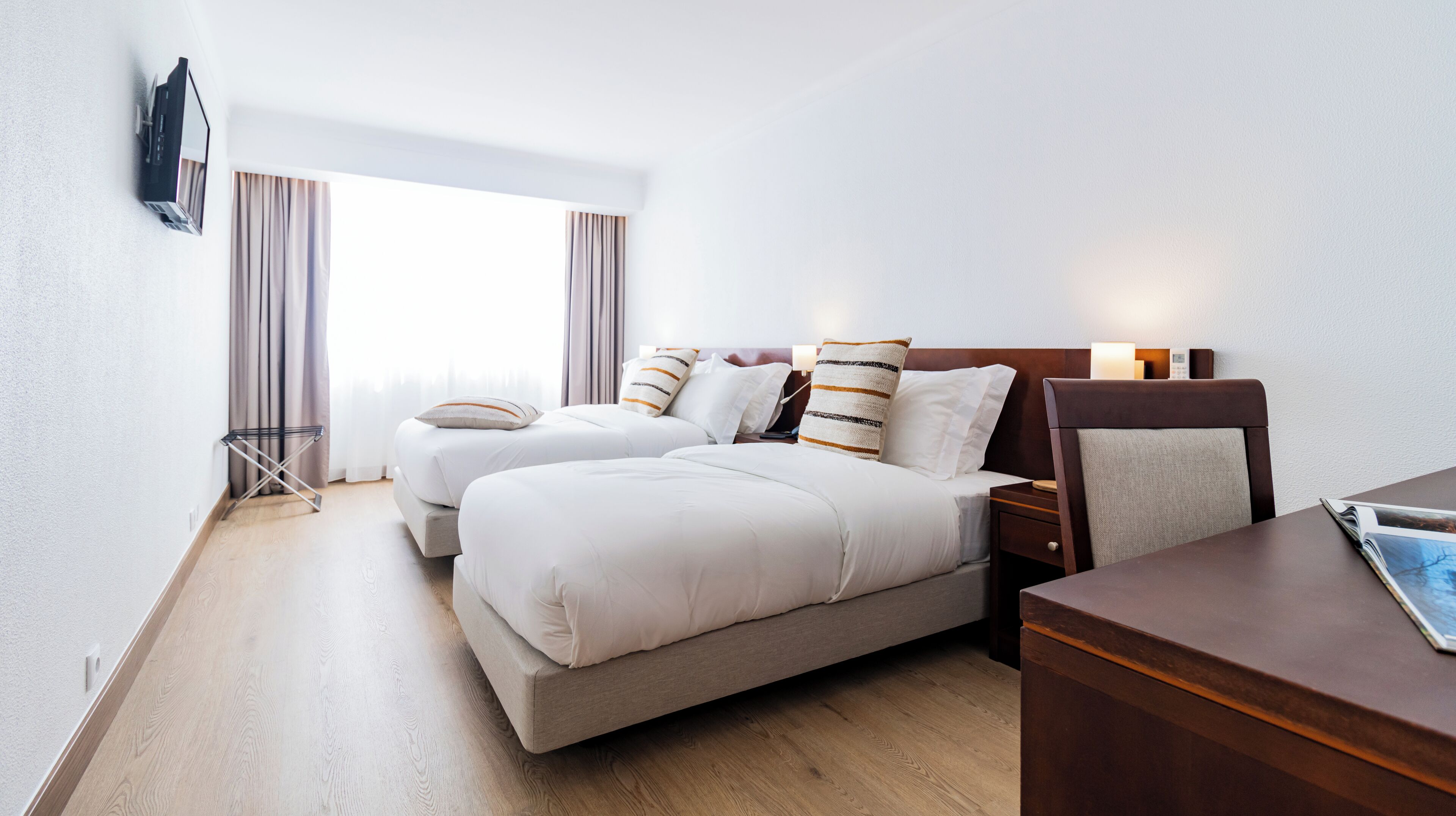 A hotel room with two beds, a desk, a TV mounted on the wall, and a window with curtains.