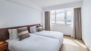 A hotel room with two beds, a wooden headboard, a nightstand, a lamp, and a view of the ocean through a window with curtains.