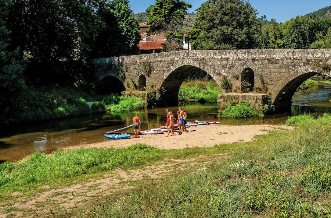 Eine Steinbrücke über einem Fluss, mit Personen und Booten am Ufer.