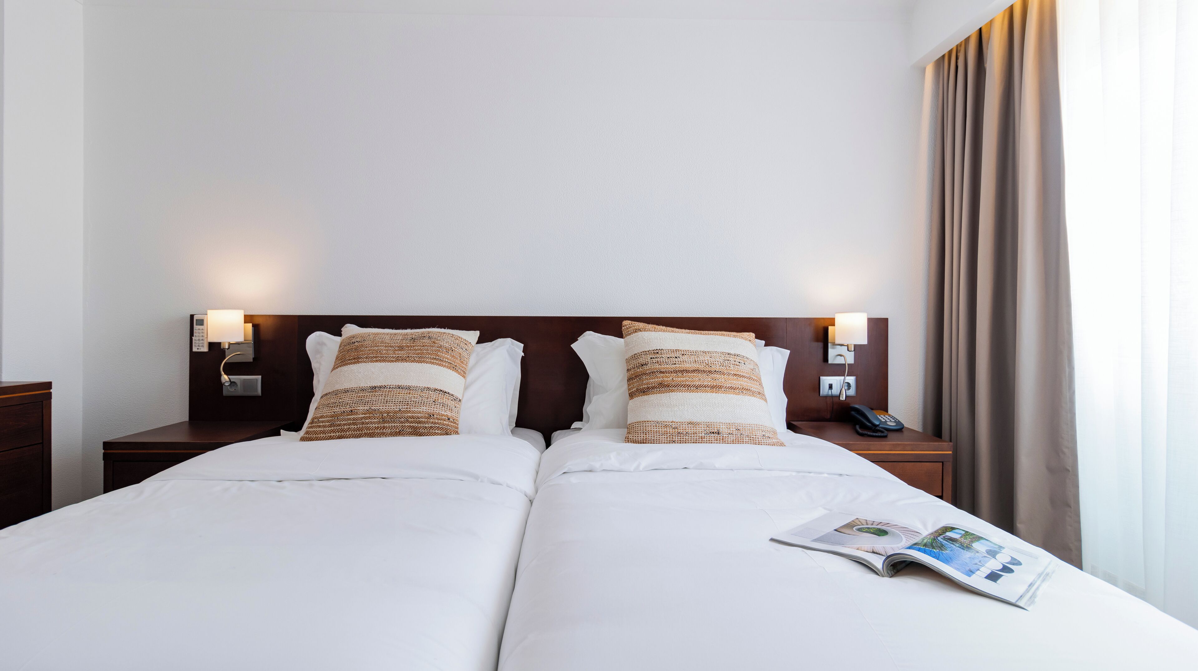 A double bed with white linens, two decorative pillows, a bedside table with a lamp, and a magazine on the bed.