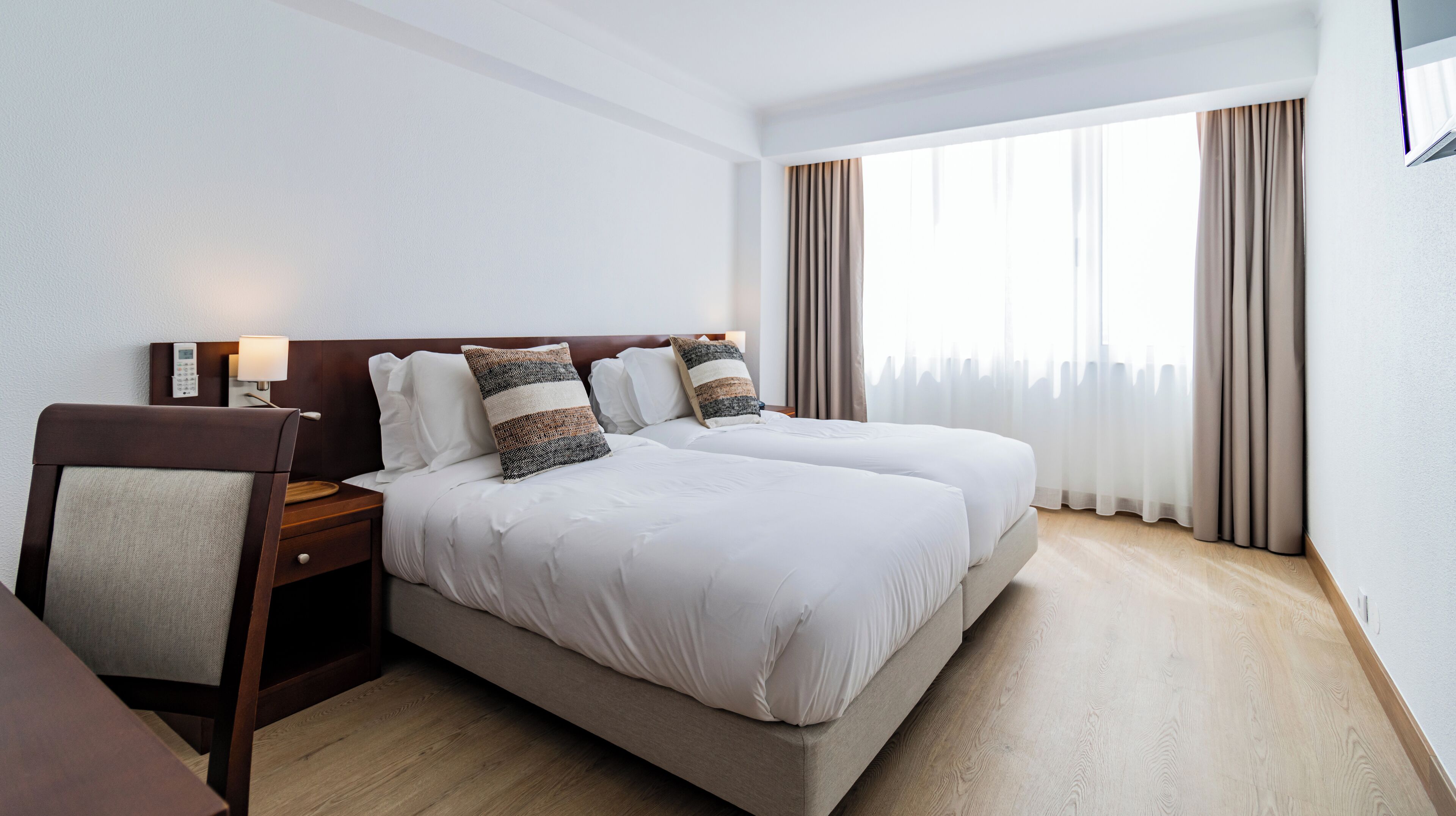 A hotel room with two beds, a desk, a chair, a bedside table with a lamp, and a large window with curtains.