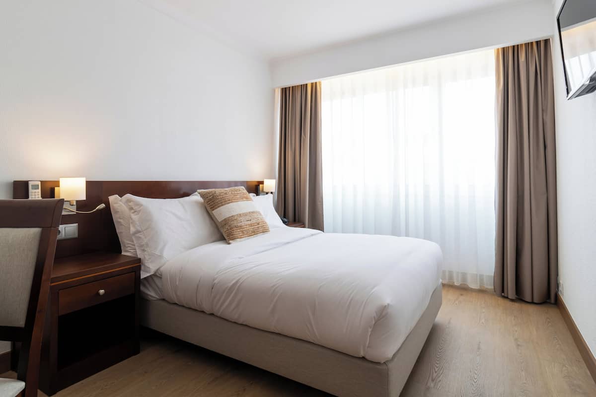 Una habitació d'hotel amb un llit, una taula, una cadira, un televisor fixat a la paret i una finestra amb cortines.