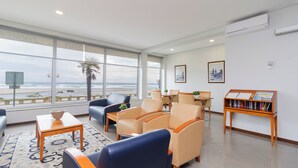 A hotel room with a large window offering a view of the beach, a seating area with chairs and a table, and a bookshelf with travel books.
