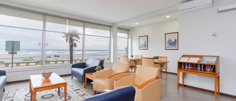 Une chambre d’hôtel dotée d’une grande fenêtre donnant sur la plage, d’un coin salon avec des fauteuils et une table, ainsi que d’une bibliothèque contenant des ouvrages de voyage.