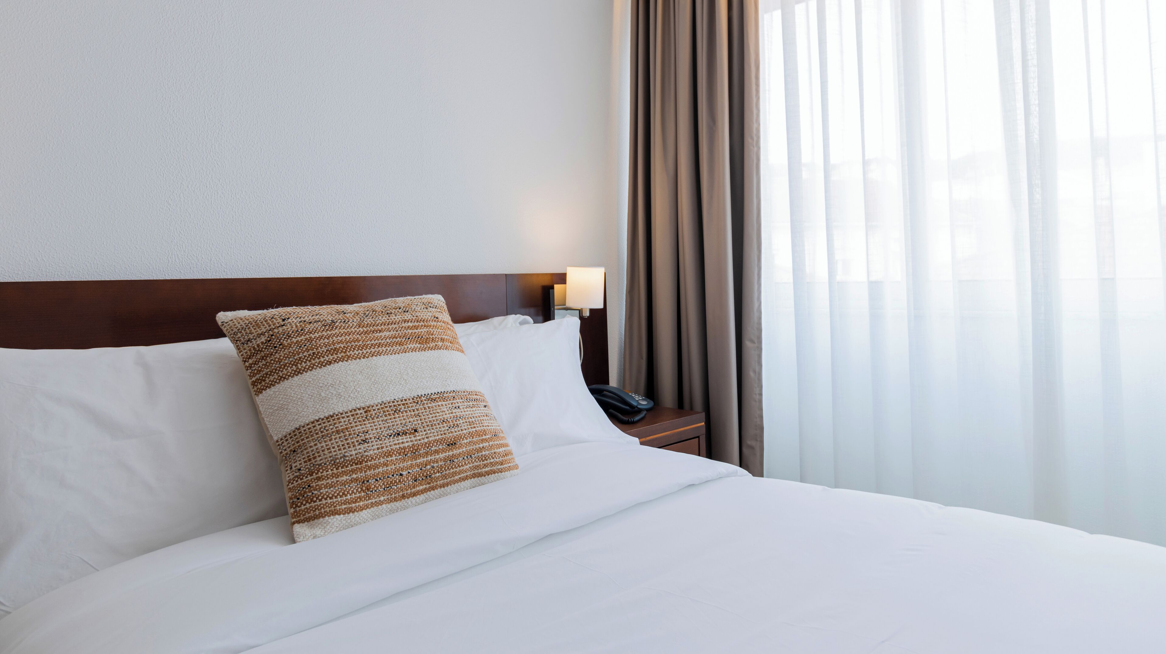 A hotel room with a neatly made bed, a wooden headboard, a bedside lamp, and a view of a building through sheer curtains.