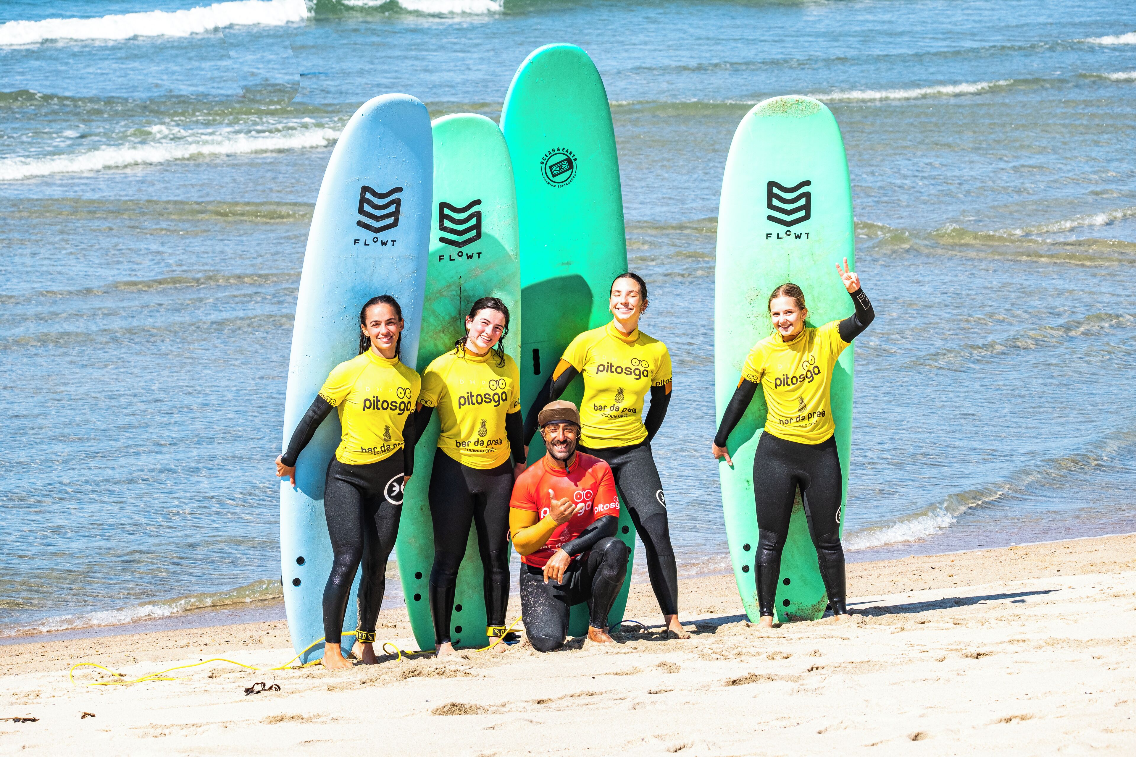 Un grupo de personas en una playa con tablas de surf, usando camisetas amarillas con logotipos, y el océano al fondo.