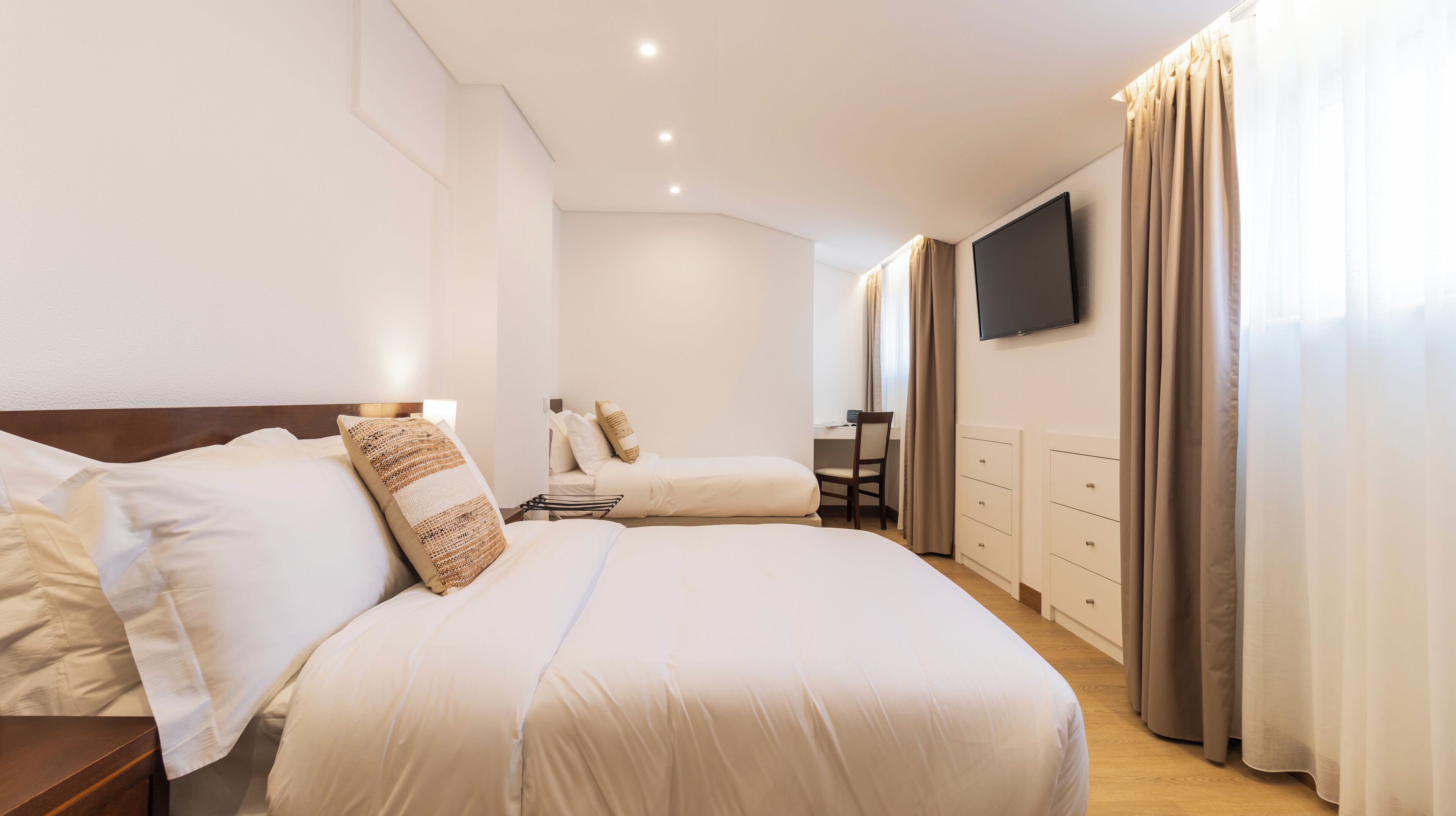 A hotel room with a large bed, a smaller bed, a desk, a chair, a TV, and a wardrobe.