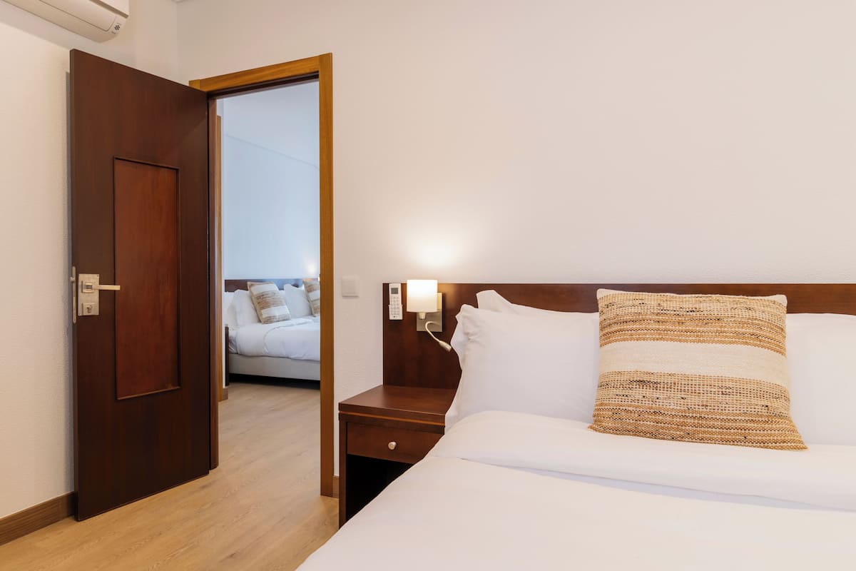 Habitació d'hotel amb una porta de fusta, un llit amb roba de llit blanca i un coixí teixit, una taula de nit amb una làmpada, i vista a una altra habitació per una porta oberta.