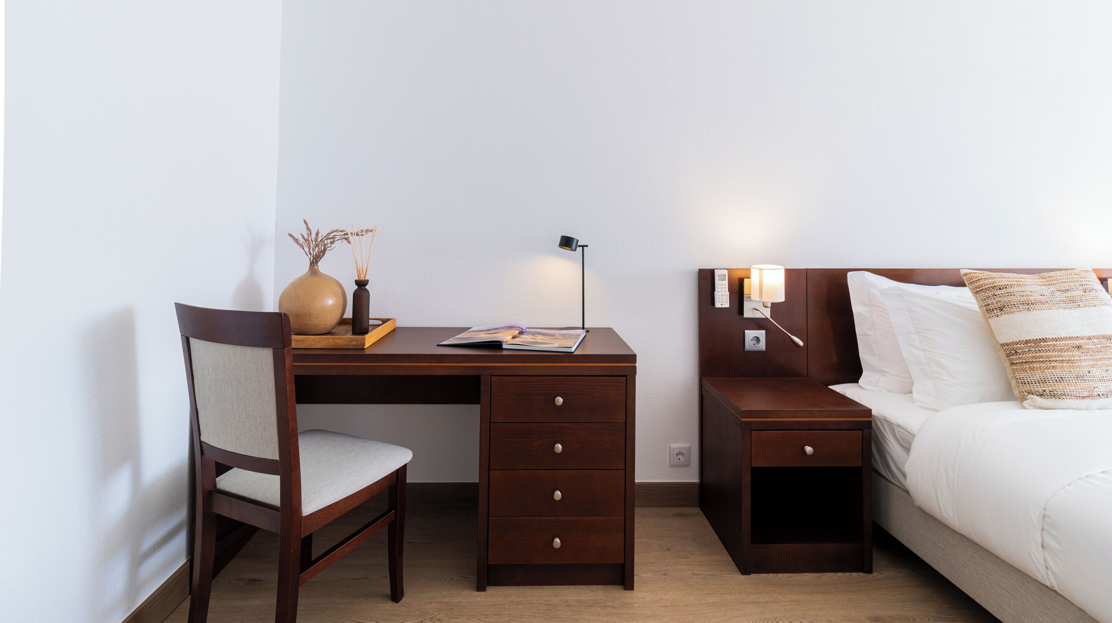 A hotel room with a wooden desk, a chair, a bedside table with a lamp, a bed with white bedding, and a nightstand with a phone and lamp.