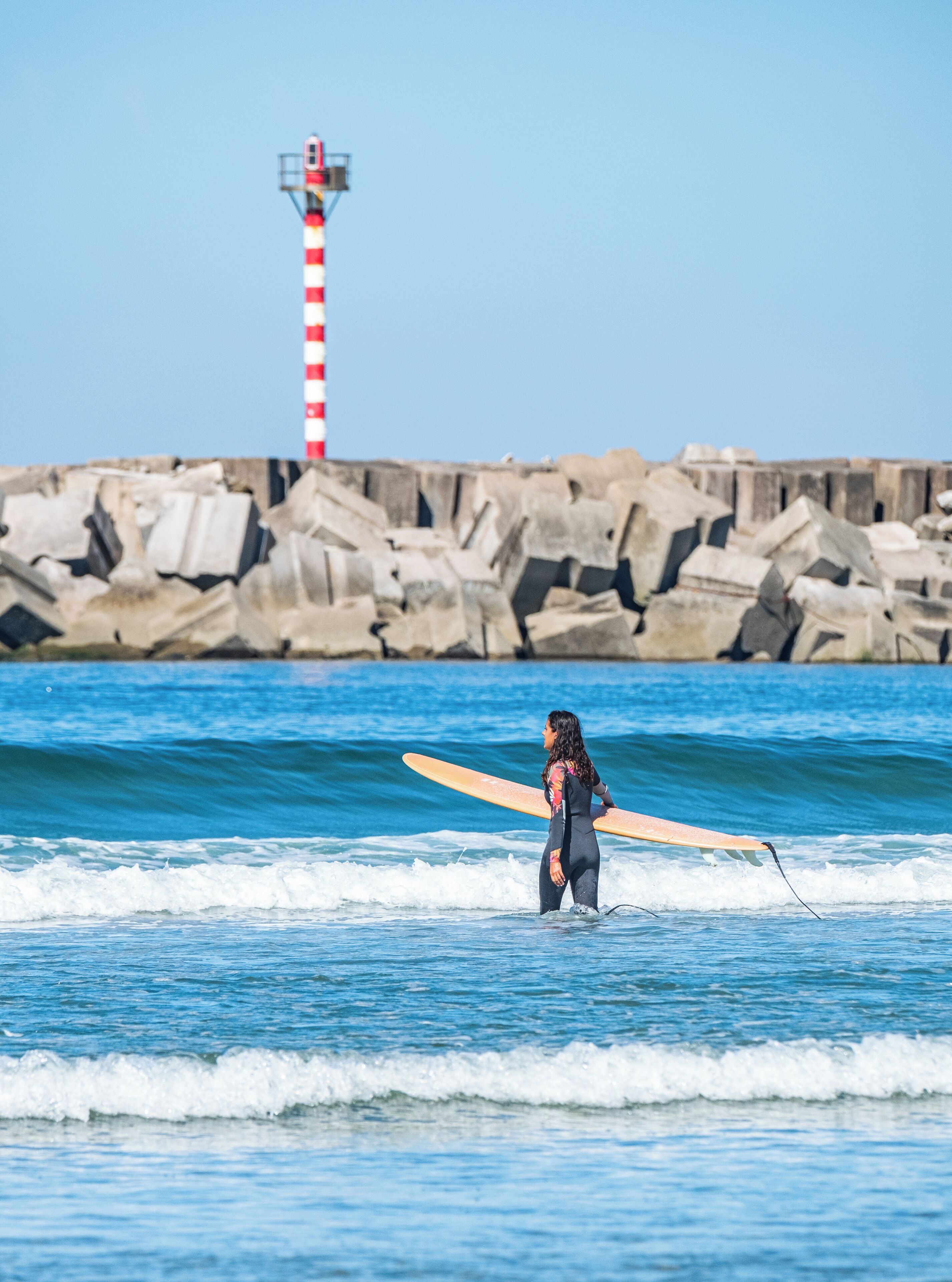 Una persona con una muta che porta un'asse da surf vicino a un molo roccioso con un faro.