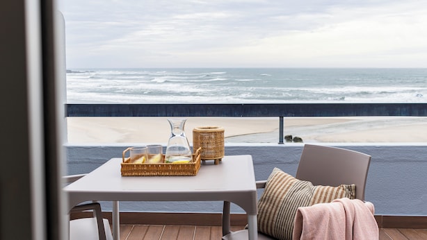 A balcony with a table set for two, overlooking a beach with waves.