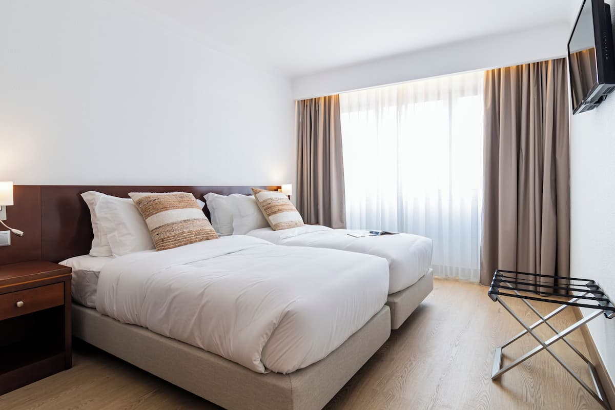Una habitació d'hotel amb dos llits, una televisió penjada a la paret, una taula de nit amb una làmpada, i una finestra amb cortines.