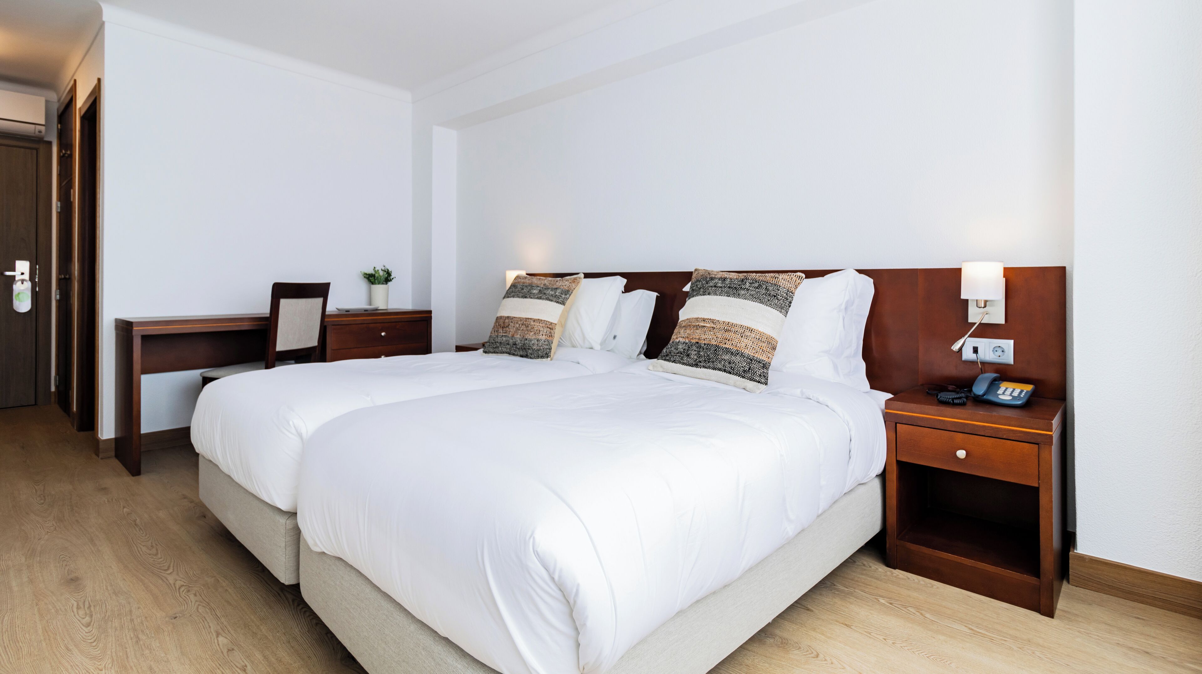 A hotel room with a large bed, a desk, a chair, a nightstand with a phone, and a wall-mounted light.