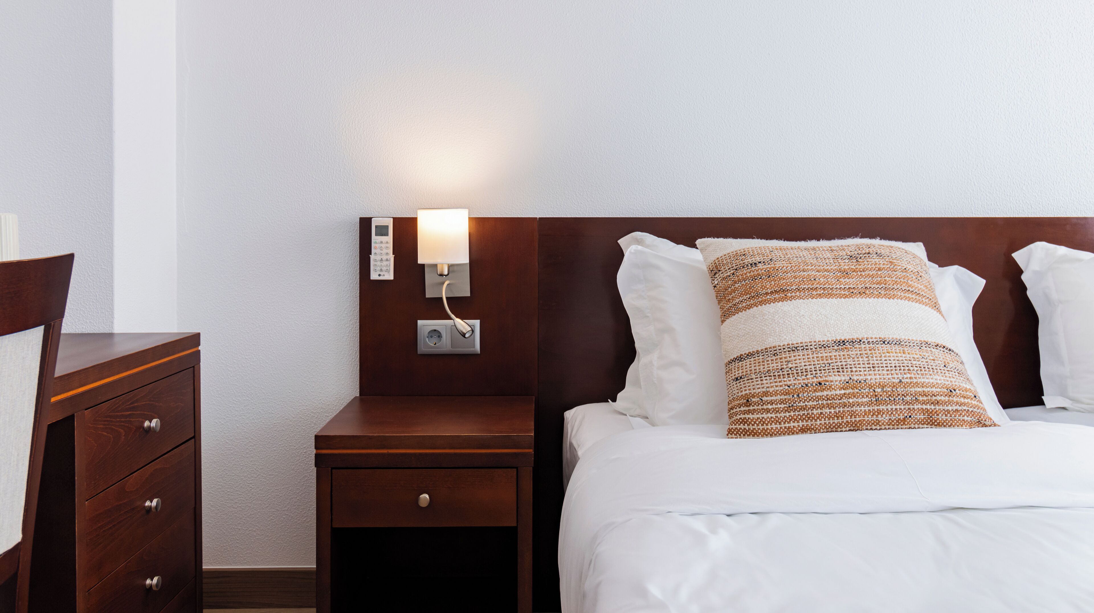 A hotel room with a wooden nightstand, a bedside table, a modern wall-mounted thermostat, and a bed with white bedding and a textured pillow.