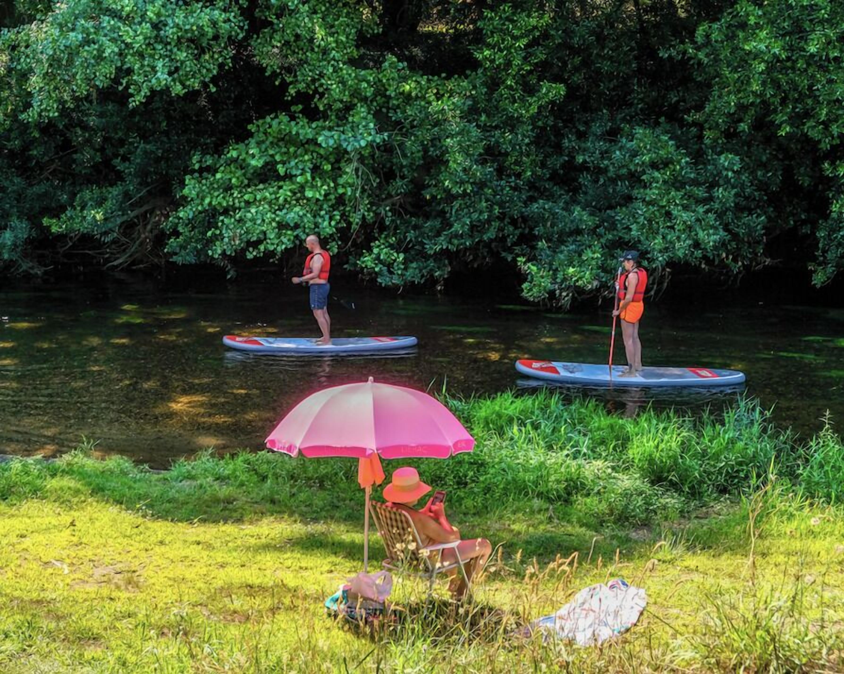 Dos personas haciendo paddleboard en un río, con una persona sentada en una silla bajo un paraguas rosa.