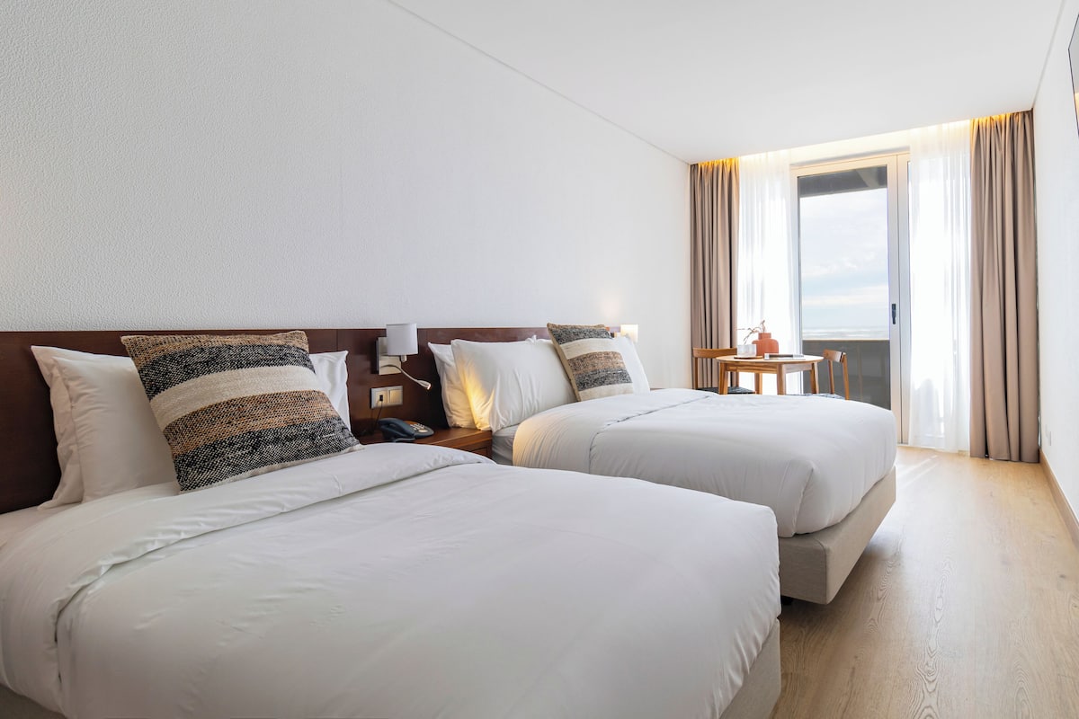 Habitació d'hotel amb dos llits, una taula de nit amb una làmpada, una taula petita amb una cadira i vistes a l'exterior a través d'una finestra.