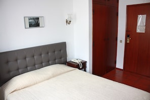 Une chambre à coucher avec une tête de lit recouverte d’un tissu gris, une couvre-lit blanche, une table de chevet avec un téléphone, un tableau encadré accroché au mur et une porte en bois sur laquelle est apposée une affiche.