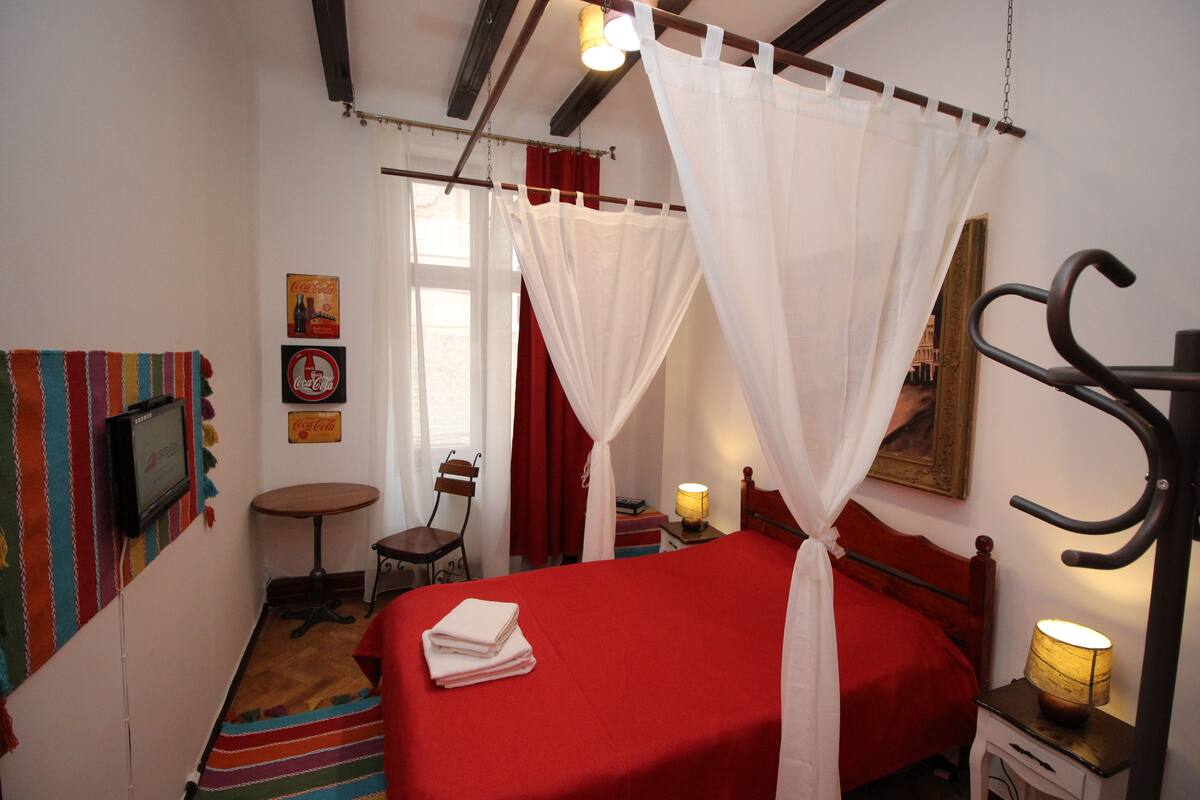 Um quarto com uma cama, uma cadeira, uma mesinha, uma luminária e uma televisão.