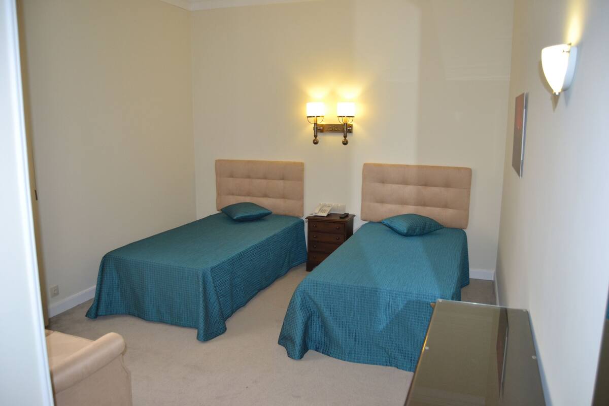 A hotel room with two single beds, a nightstand, a telephone, and wall-mounted lighting.