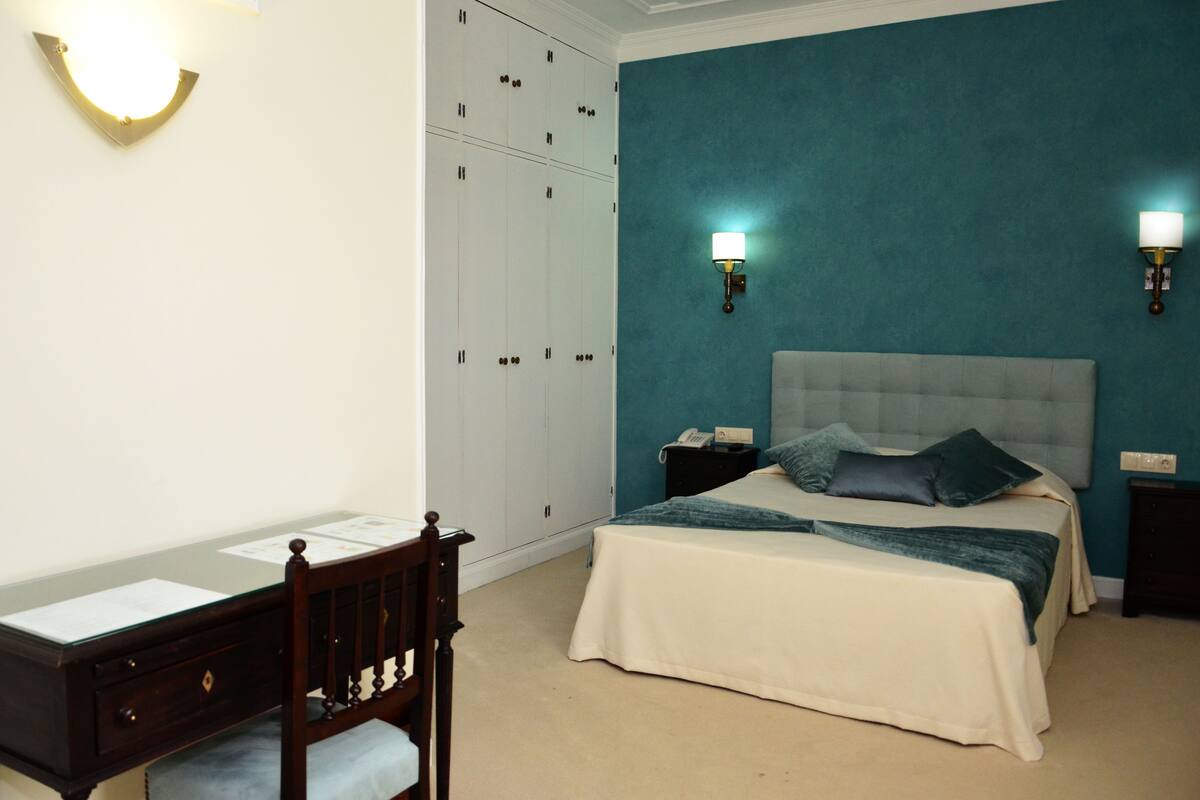 A bedroom with a bed, a desk, and a chair. There are two wall sconces and a wardrobe.