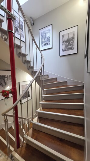 Una escalera con barandillas blancas y peldaños de madera, flanqueada por tuberías rojas y enmarcada por fotografías blanco y negro en las paredes.
