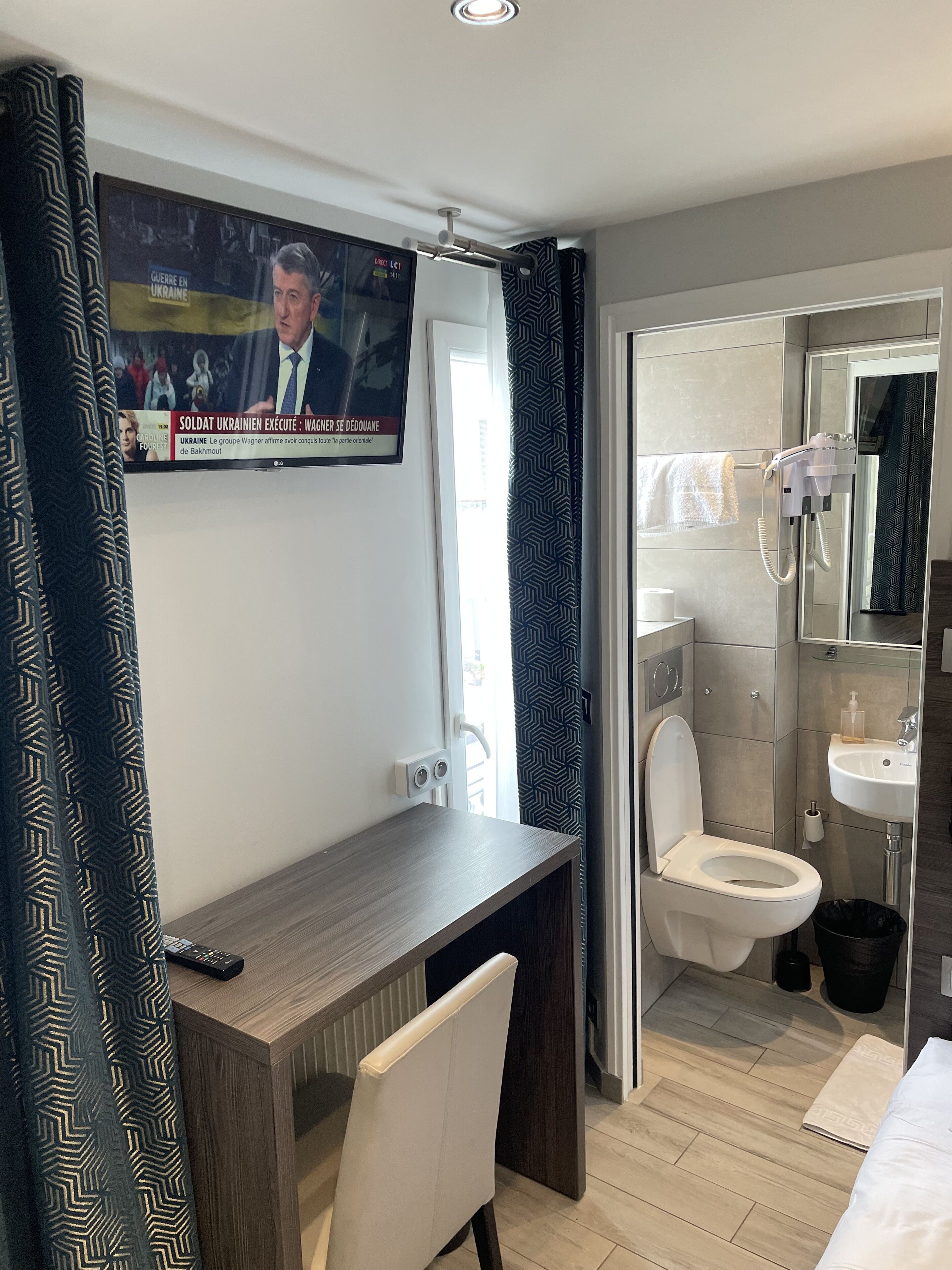 Una habitación de hotel moderna con un escritorio de madera, un televisor montado en la pared, y un baño con inodoro, lavabo y espejo.