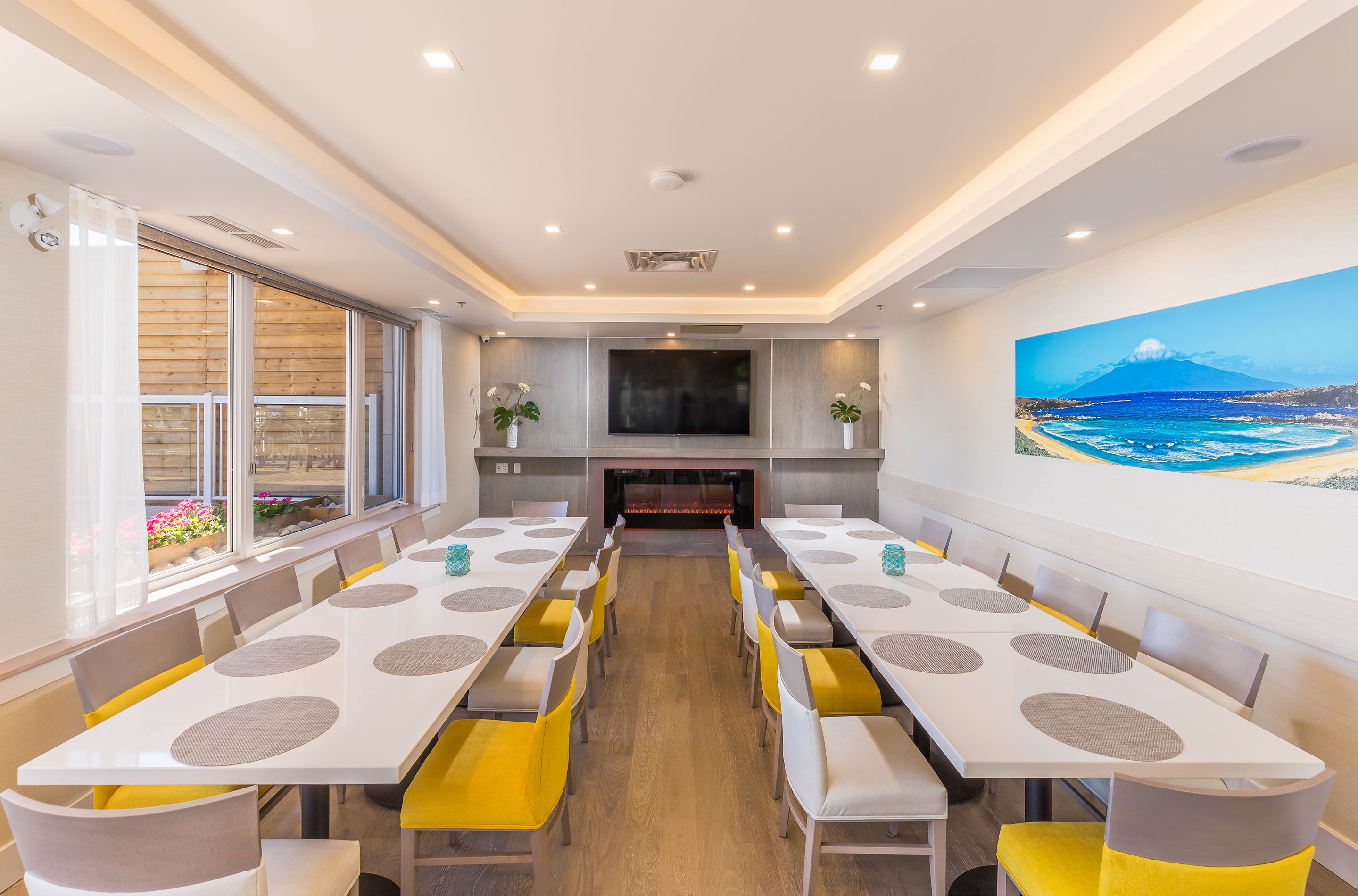 A spacious dining area with long tables and yellow chairs, a large TV screen, and a painting of a beach scene on the wall.